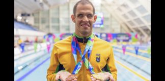 Vestindo a jaqueta amarela do Brasil, com a as piscinas ao fundo, Matheus Rheine exibe a medalha de bronze na mão direita e a de ouro na mão esquerda, no Mundial de Natação Paralímpica em Manchester, 2023